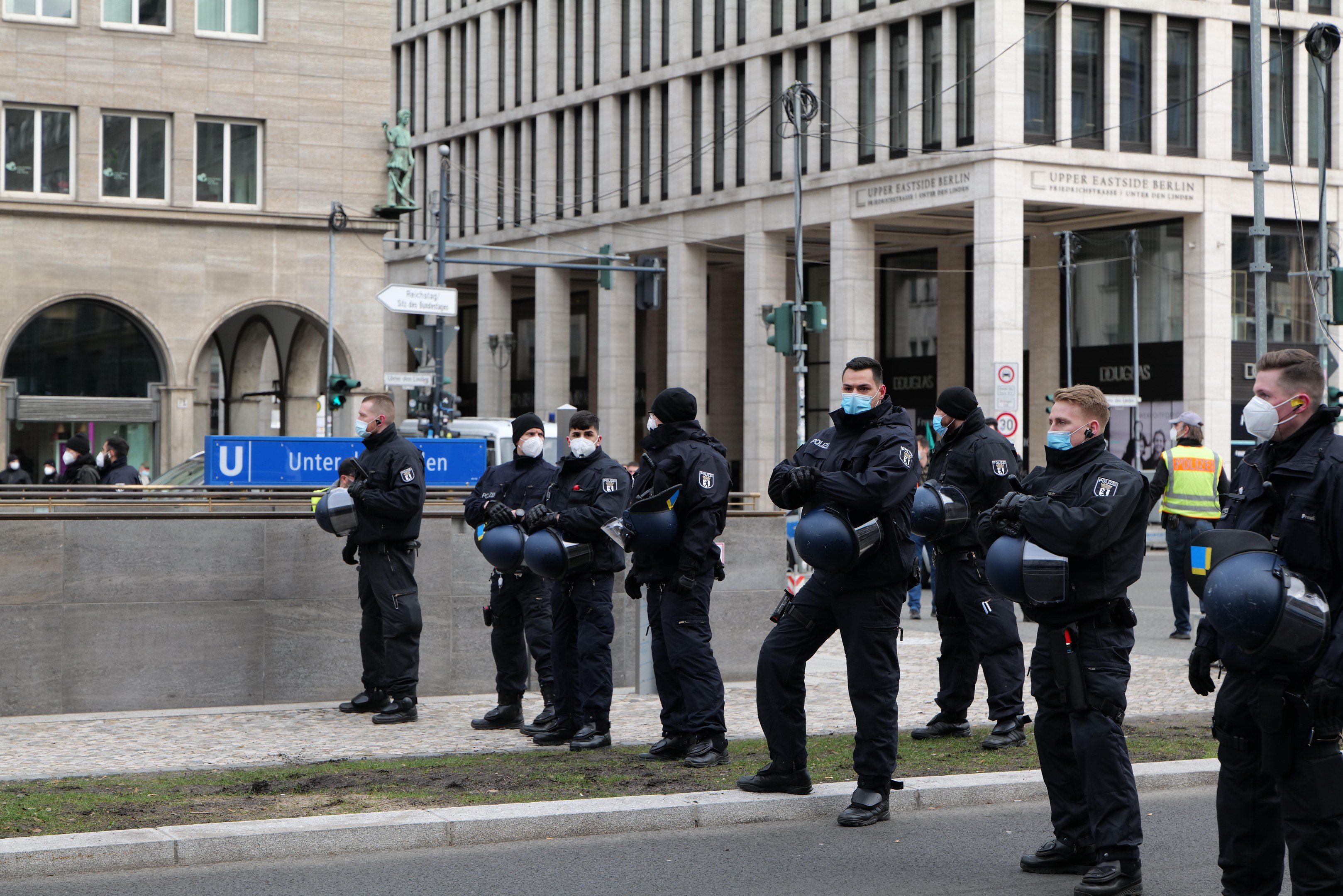 Eine Gruppe von Polizisten in schwarzen Uniformen und Masken steht vor einem Berliner Gebäude mit Glasfenstern und Säulen, einige halten Helme, mit Laternen, Verkehrszeichen, Schildern mit Text, einer Statue und anderen Menschen im Hintergrund.