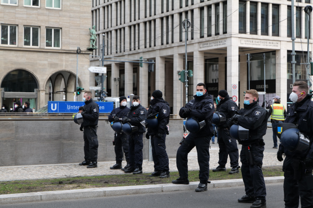 Eine Gruppe von Polizisten in schwarzen Uniformen und Masken steht vor einem Berliner Gebäude mit Glasfenstern und Säulen, einige halten Helme, mit Laternen, Verkehrszeichen, Schildern mit Text, einer Statue und anderen Menschen im Hintergrund.