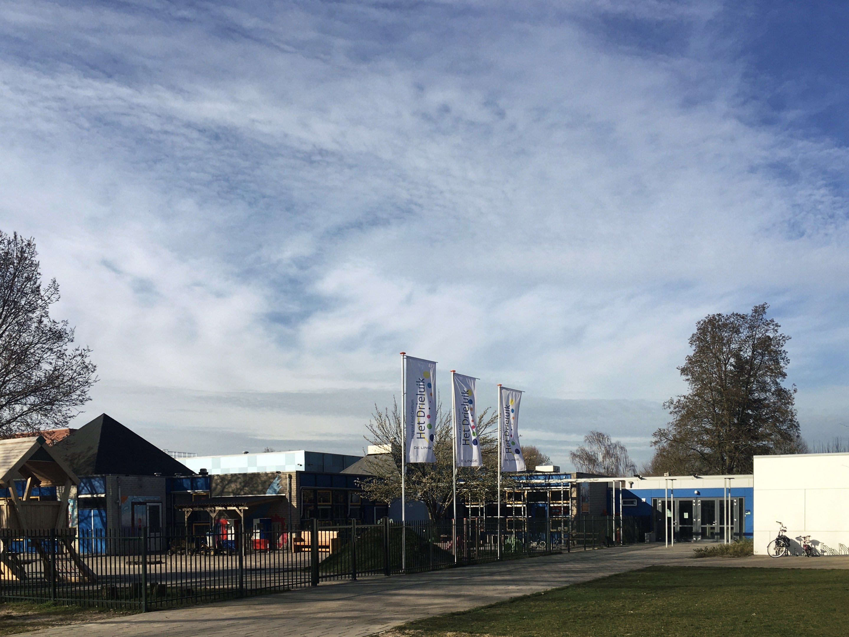 Großes Gebäude mit einem grasbewachsenen Bereich davor, umgeben von einem Metallzaun, mit einem Weg, Bäumen, Pfosten, Bannern, einem geparkten Fahrrad, nahen Häusern und einem bewölkten Himmel, identifiziert als der neue Spielplatz der Schule.