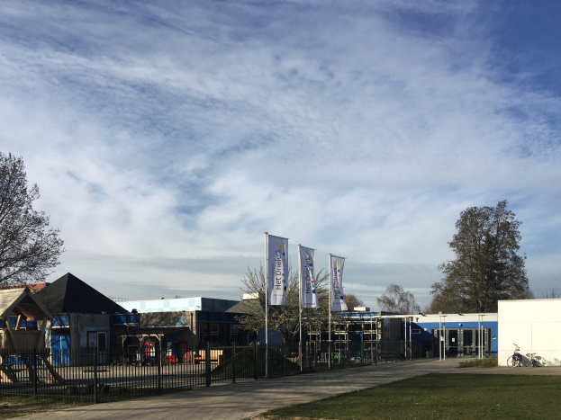 Großes Gebäude mit einem grasbewachsenen Bereich davor, umgeben von einem Metallzaun, mit einem Weg, Bäumen, Pfosten, Bannern, einem geparkten Fahrrad, nahen Häusern und einem bewölkten Himmel, identifiziert als der neue Spielplatz der Schule.
