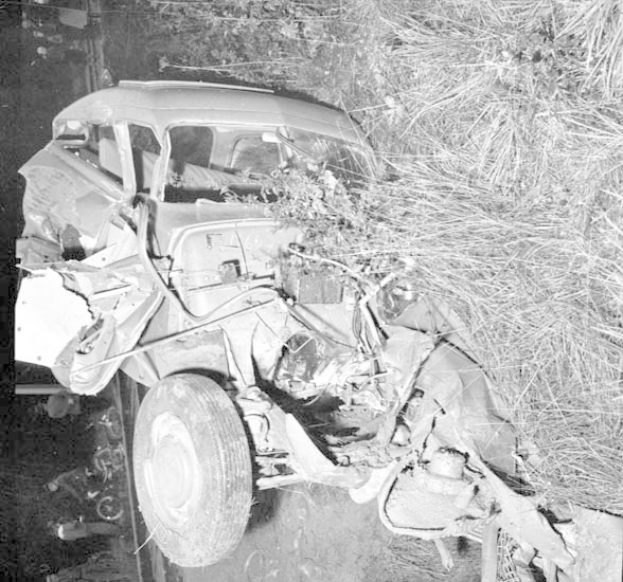 Schwarz-weißes Foto eines stark beschädigten Autos am Straßenrand, mit eingedellter Front, zersplitterten Fenstern, einem Baum im Hintergrund und verstreuter Trümmer.