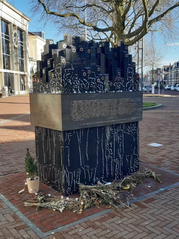 Denkmal für Holocaust-Opfer in einem Stadtplatz mit einem Baum davor, Topfpflanze, verstreuten trockenen Blättern und Gebäuden, Fahrzeugen, Pfählen und Bäumen im Hintergrund.