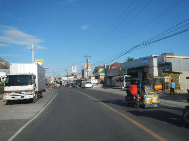Stadtstraße mit einem Lkw, der an Gebäuden, Strommästen mit Drähten, Schildern, Bäumen und einem bewölktem Himmel vorbeifährt.