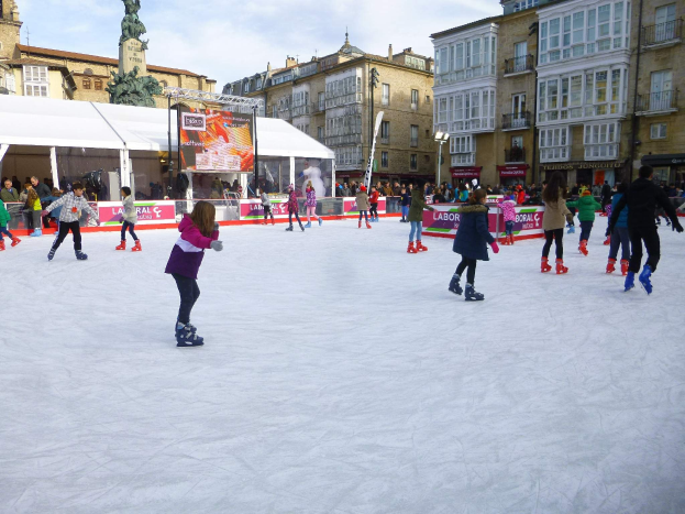 Menschen, die auf einer Outdoor-Eisbahn in einer städtischen Umgebung mit Gebäuden, Laternenmasten, einer Statue, Bannern und Zelten im Hintergrund Schlittschuh laufen.