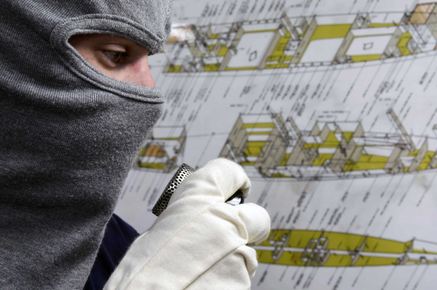 Ein Mann mit Skimaske und Handschuhen hält ein Objekt, während er vor einer Wand mit einem Blaupause steht.