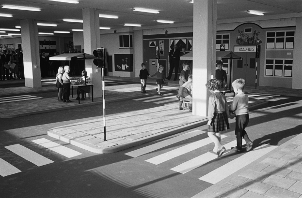 Schwarz-weiß-Foto von Kindern, die eine Straße an einer Ampel überqueren, mit Fußgängern in der Nähe, einer Verkehrsampel im Vordergrund und strukturellen Säulen mit Leuchten im Hintergrund.