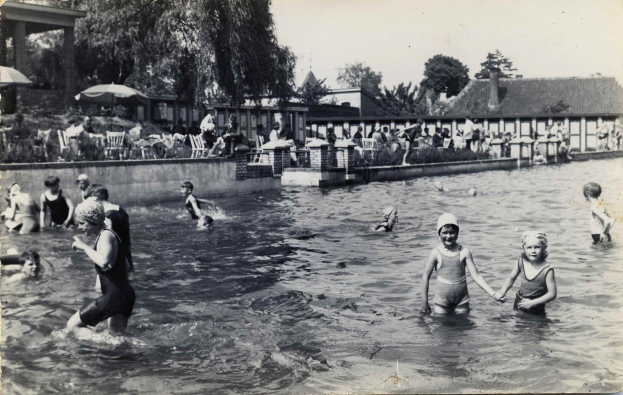 Schwarzes und weißes Foto von Kindern, die in einem Swimmingpool spielen, mit Menschen im Hintergrund unter Schirmen, in der Nähe von Gebäuden und Bäumen unter einem klaren Himmel.