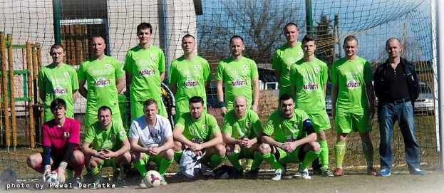 Eine Gruppe von Männern in grünen T-Shirts steht auf einem Fußballfeld, einige in Hockstellung, mit einem Ball vor ihnen neben einem Torpfosten, Bäume, Gebäude und Fahrzeuge im Hintergrund.
