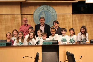 Eine Gruppe von Kindern und Erwachsenen steht lachend vor einem Rednerpult, mit Monitoren, Mikrofonen und anderen Gegenständen auf einem Tisch im Vordergrund, einem Logo an der Wand und einem Bildschirm auf der rechten Seite, was an eine Stadtratssitzung erinnert.