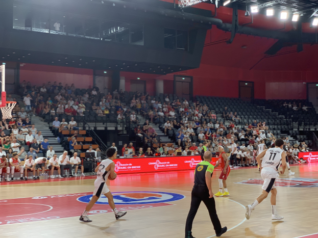 Eine Gruppe von Menschen, die Basketball in einer Turnhalle mit einem Netz auf der linken Seite und Zuschauern im Hintergrund unter Stadionbeleuchtung spielt.