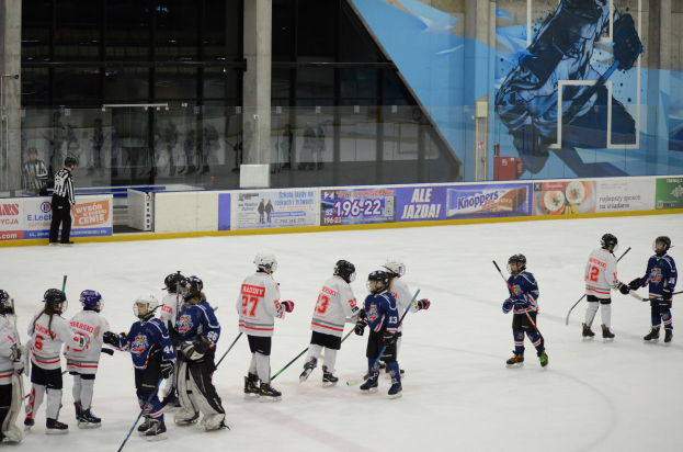 Eine Gruppe junger Menschen, die auf einer Indoor-Eisfläche Hockey spielen, tragen Helme und halten Stöcke in der Hand, mit einer Person links daneben und Bannern, Säulen und Glaswänden im Hintergrund.