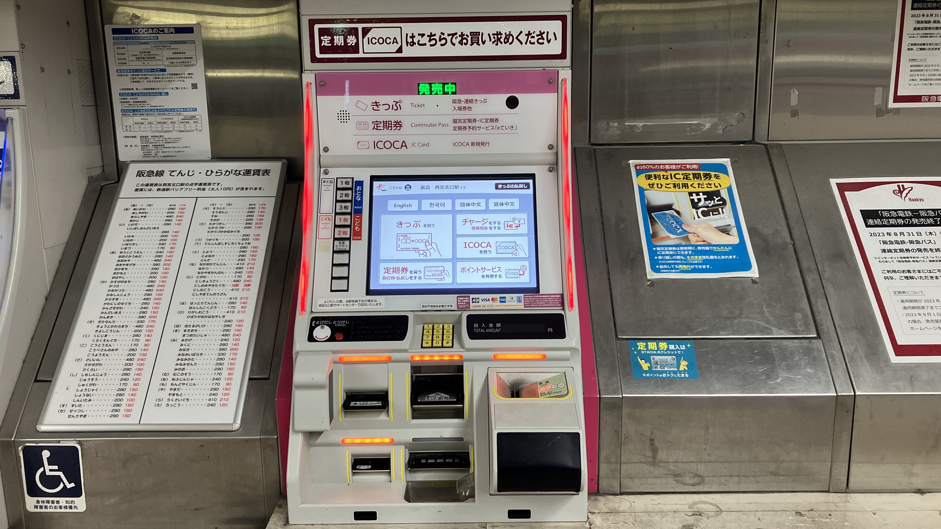 Geldautomat in einer U-Bahn-Station mit einem Bildschirm, Tasten und Plakaten an der Wand dahinter.