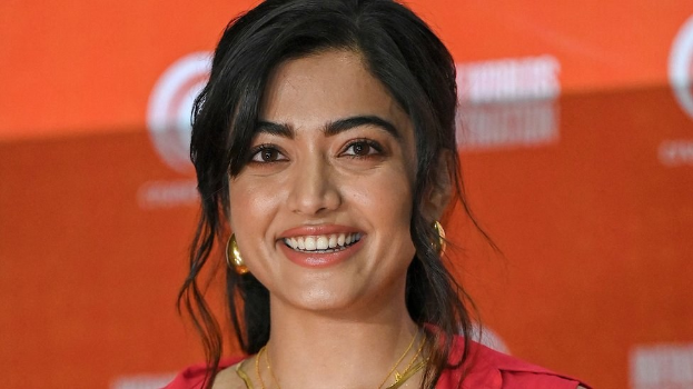 Eine Frau in einem roten Kleid mit einer glatten Hochsteckfrisur und dezentem Make-up lächelt in die Kamera, trägt schwarze High Heels und hat ein hellgelbes Banner mit der Aufschrift 'Die besten Schönheits Looks von Sonam Kapoor beim Cannes Film Festival' im Hintergrund.