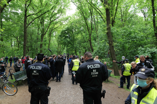 Polizeibeamte vor einer Menschenmenge bei einer Outdoor-Veranstaltung mit Fahrrädern und einer Bank im Vordergrund und Bäumen im Hintergrund.