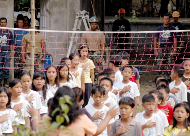 Eine Gruppe von Kindern steht vor einem Netz mit einer Frau im Vordergrund, während im Hintergrund einige Menschen mit Helmen, eine Leiter, eine Wand und Bäume zu sehen sind.