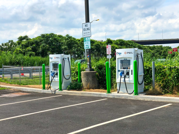 Ladestation für Elektrofahrzeuge auf einem Parkplatz mit umgebenen Infrastruktur, Grünflächen und einer Brücke im Hintergrund.