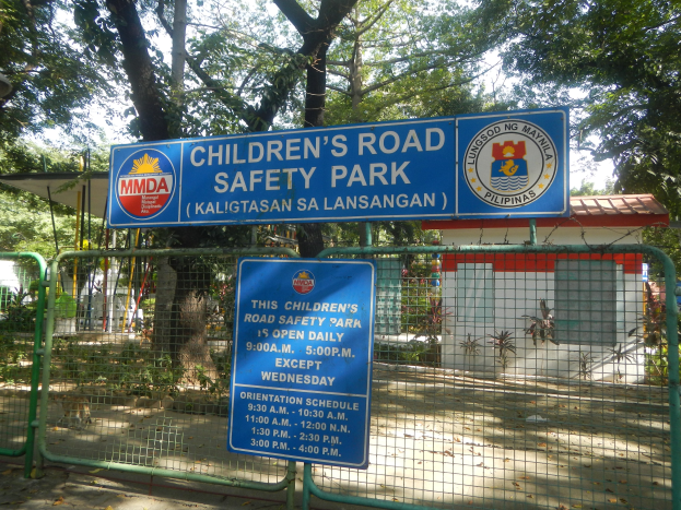 Metallzaun, der einen Kindersicherheitspark für Straßenverkehr umgibt, mit zwei blauen Informationsschildern, einem kleinen Haus mit Dach, Pflanzen, Bäumen und einem bewölkten Himmel.