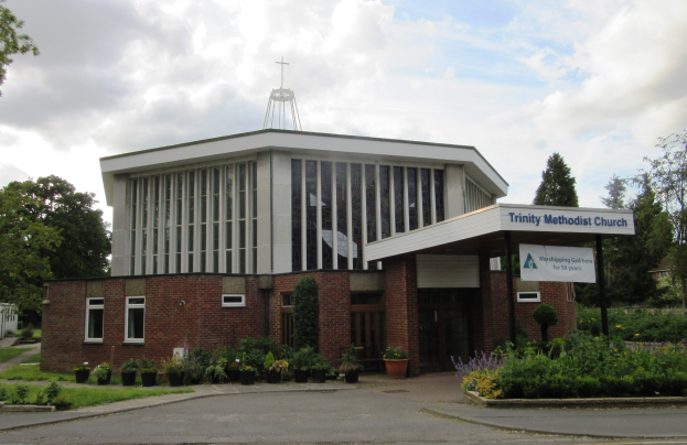 Außenansicht der Trinity Methodist Church in Stoke-on-Trent, Staffordshire, mit Topfpflanzen, Gras, Bäumen, einem Schild, einem Straßenpfahl und einem Weg unter einem bewölkten Himmel.
