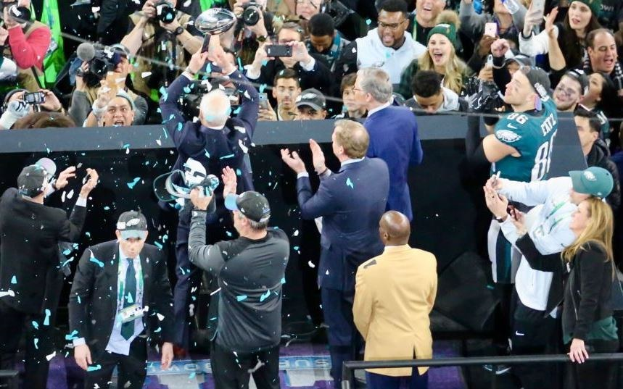 Philadelphia Eagles Spieler feiern mit dem Vince Lombardi Trophy nach dem Sieg im Super Bowl, einige tragen Mützen und halten den Pokal, während andere Konfetti werfen, mit einem Geländer und Fotografen im Hintergrund.