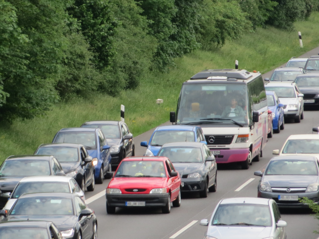 Ein Stau auf einer Autobahn mit vielen Autos, einem Lieferwagen und Menschen in den Fahrzeugen, mit Bäumen und Gras im Hintergrund.