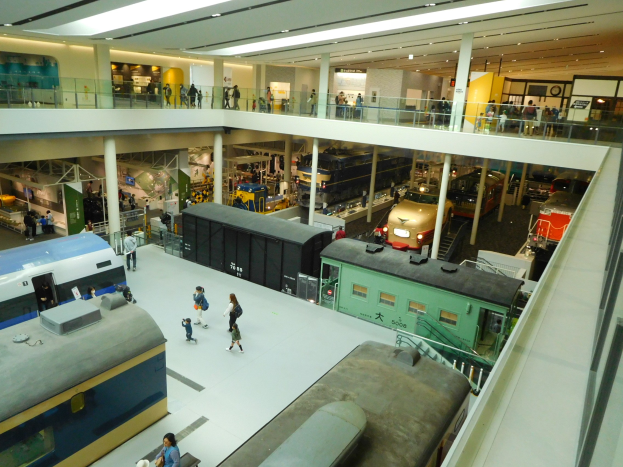 Ein gro├če Museumshalle mit verschiedenen Zugtypen und Menschen, die beobachten, Gel├äder, Pfeiler, Deckenleuchten und Informationsschilder.