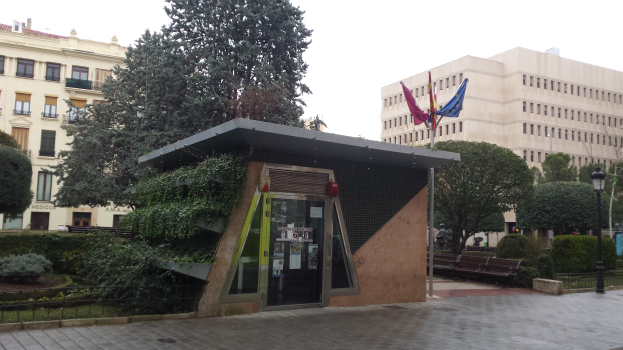 Kleines grünes Dach auf einem Gebäude in einer städtischen Parkanlage mit umliegenden Bäumen, Bänken, Laternen und Fahnen.