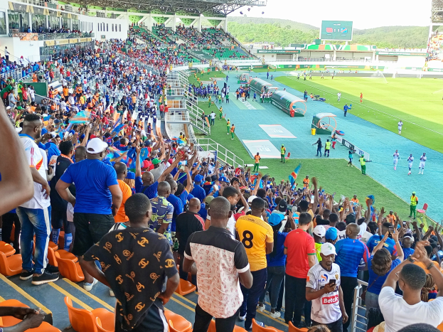 Eine große Menschenmenge versammelt sich vor einem eingezäunten Fußballfeld unter einem klaren blauen Himmel, mit Hügeln im Hintergrund, einige sitzen auf Stühlen und andere stehen, in Erwartung des Beginns eines Spiels.