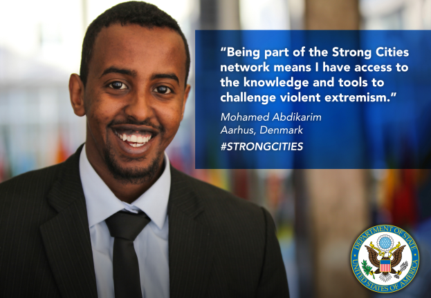 Ein Mann im Anzug und Krawatte lächelt in die Kamera, mit einem Logo und Text auf der rechten Seite, der besagt: "Teil des starken Städtenetzwerks zu sein bedeutet, Zugang zu Wissen und Werkzeugen zu haben, um gewalttätigen Extremismus in Frage zu stellen", und einem unscharfen Hintergrund.