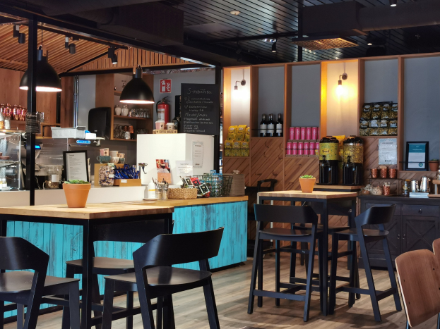 Innenraum eines Restaurants mit Holztischen, Stühlen, einer Topfpflanze auf einem Tisch, einer Texttafel, einem Kühlschrank, einem Feuerlöscher, Regalen mit Flaschen, Schränken mit Gegenständen, einem gerahmten Foto an der Wand und Deckenleuchten.