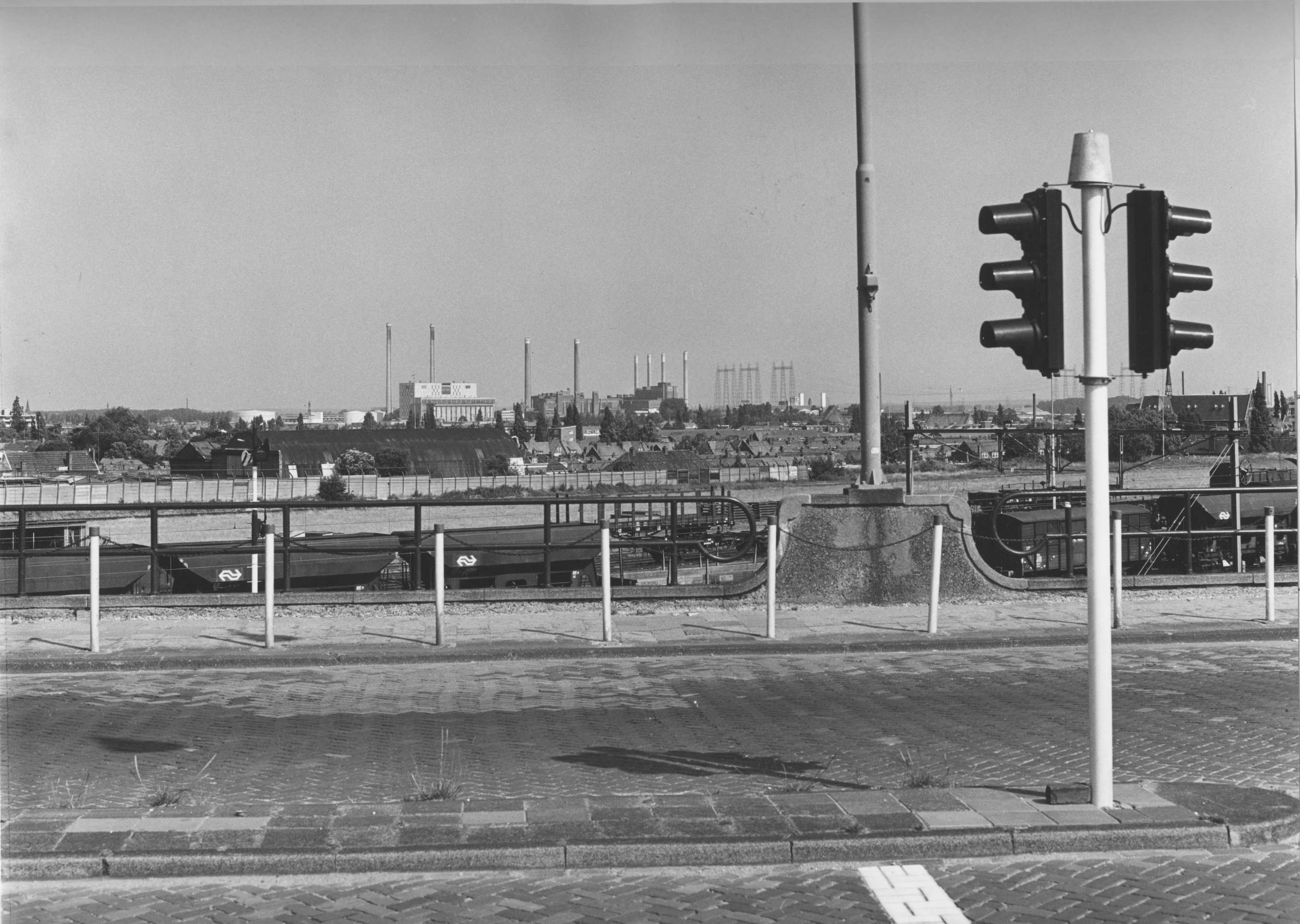 Schwarzes und weißes Foto einer Ampel an der Seite einer Straße mit Masten, einem Zaun, Bäumen, Gebäuden und einem bewölkten Himmel.