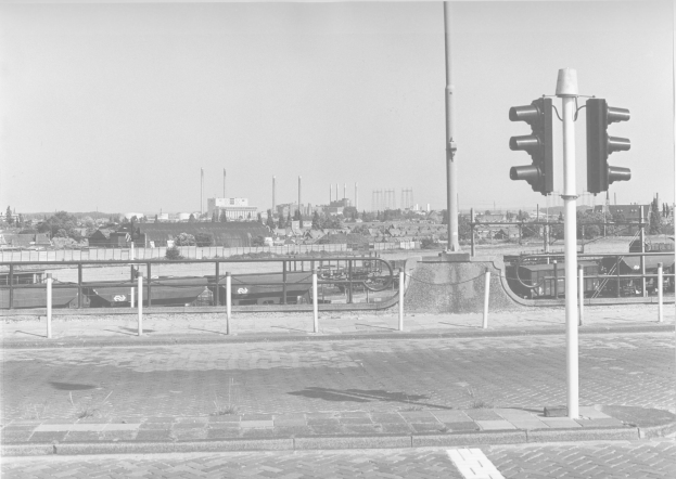 Schwarzes und weißes Foto einer Ampel an der Seite einer Straße mit Masten, einem Zaun, Bäumen, Gebäuden und einem bewölkten Himmel.