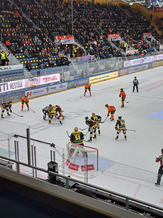 Eishockeyspieler konkurrieren auf einem Eisstadion mit einem Tor und einer Umzäunung, während Zuschauer von Stühlen aus zusehen und Banner im Hintergrund hängen, während das Stadion von Scheinwerfern beleuchtet wird.