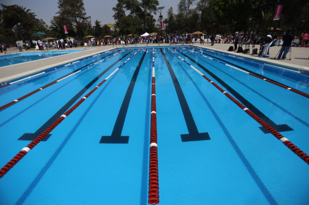 Großes Schwimmbad mit Bahnentrennern, umgeben von Menschen mit Taschen und Gegenständen, mit Zelten, Transparenten, Stangen, einem Schild und einem bewölkten Himmel im Hintergrund.