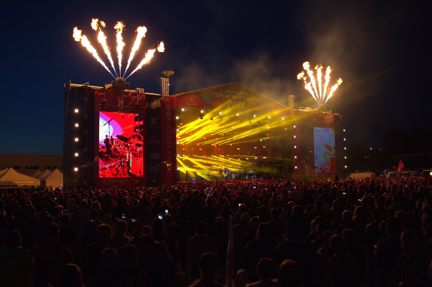 Eine große Menge bei einem Musikfestival schaut auf einer hell erleuchteten Bühne mit Bannern, Zelten links und Feuerwerk am dunklen Himmel.