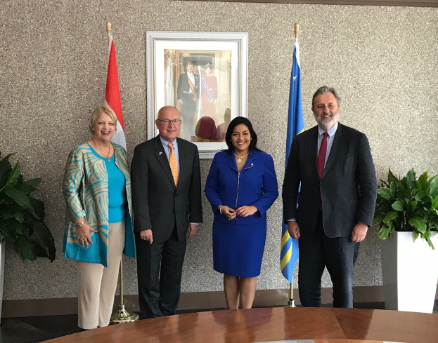 Gruppe von Menschen, die um einen Tisch mit Papieren stehen, umgeben von Fahnen, Topfpflanzen und einem Rahmenfoto an der Wand.