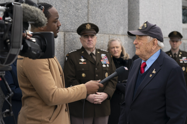 Ein Mann in Anzug und Mütze spricht in ein Mikrofon, während ein Reporter ein Objekt hält und ein Videograf im Hintergrund filmt.