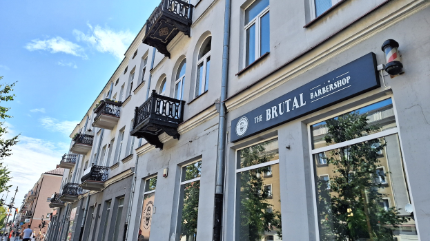 Brutalistischer Friseurladen in Berlin mit Betongebäuden, Fenstern, Geländern, Balkonen und Beschriftung, umgeben von Bäumen, Straßenlaternen und Fußgängern unter einem bewölkten Himmel.