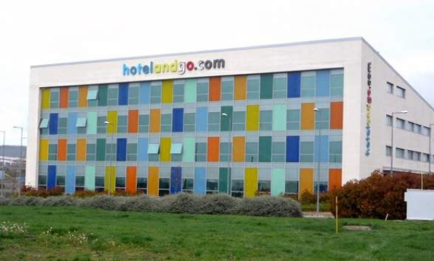 Großes modernes Hotelgebäude mit zahlreichen Fenstern, umgeben von Grünflächen, einem Zaun, Straßenlaternen, geparkten Fahrzeugen, fernen Hügeln und einem bewölkten Himmel; das Wort "hotel und go" ist auf dem Gebäude zu sehen.