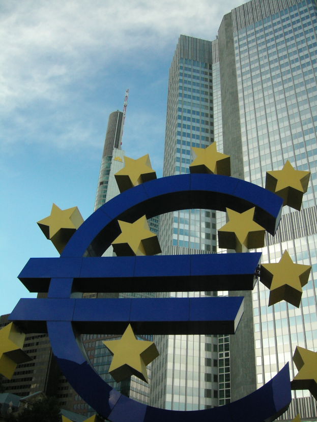 Das Euro-Zeichen steht prominent vor dem Hauptsitz der Europäischen Zentralbank, einem großen modernen Gebäude, das von Bäumen umgeben ist und unter einem bewölkten Himmel in Frankfurt, Deutschland, steht.