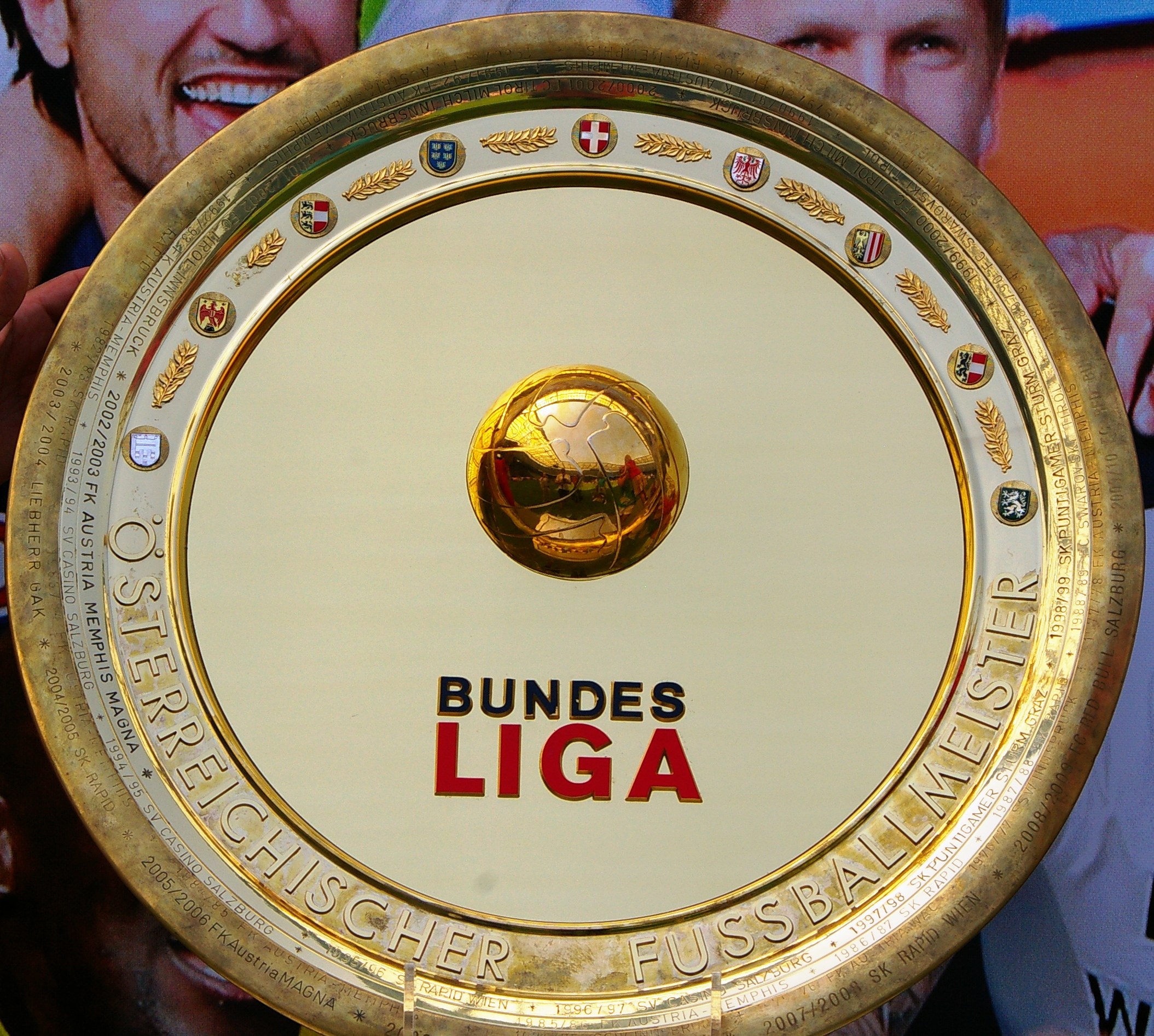 Ein Mann hält einen goldenen Schild mit der Aufschrift "Bundesliga", im Hintergrund ein Banner mit drei Personen.