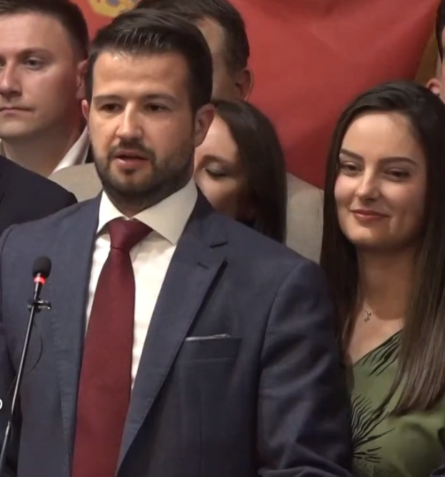 Ein Mann im Anzug steht an einem Rednerpult und spricht in ein Mikrofon, mit einem Banner im Hintergrund und einer Menge davor, wahrscheinlich wegen eines Social-Media-Verbots.