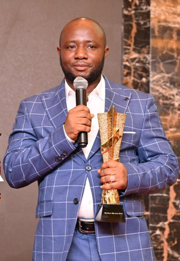 Ein Mann in einem blauen Anzug hält ein Mikrofon und eine Trophäe und steht selbstbewusst mit einem freudigen Gesichtsausdruck, wahrscheinlich feiert er einen Award-Sieg.