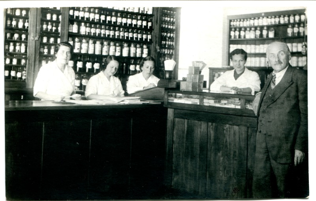 Ein Schwarz-Weiß-Foto einer Gruppe von Menschen, die vor einer Theke in einer Apotheke stehen, mit Papieren und Gegenständen auf der Theke und Regalen, die mit Flaschen gefüllt sind, im Hintergrund.