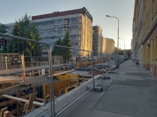 Stadtstraße mit einer Baustelle in der Mitte, umgeben von Gebäuden, Straßenlaternen, Straßenlaternen, Fahrzeugen, Absperrpoller, Bäumen und einem klaren blauen Himmel.