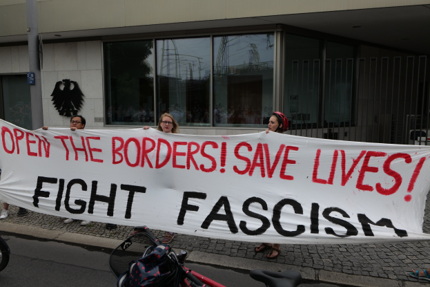 Protestierende halten ein Transparent mit der Aufschrift 'Öffnet die Grenzen, rettet Leben, kämpft gegen Faschismus' vor einem Gebäude mit Glasfenstern und einem Pfahl, mit einem Fahrrad und einer Tasche auf der Straße.