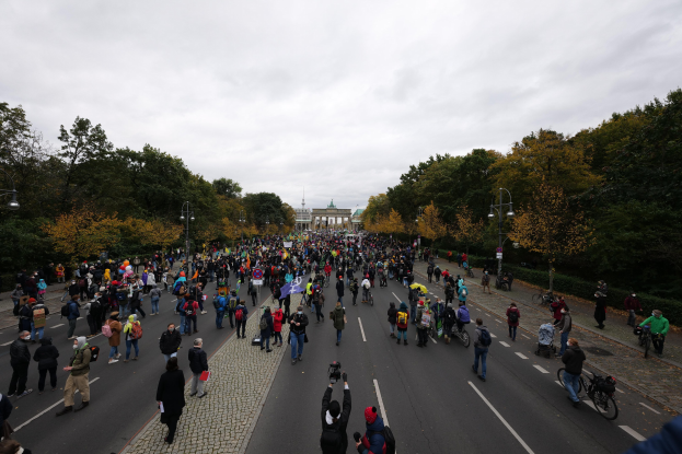 Eine große Gruppe von Menschen marschiert auf einer von Bäumen gesäumten Straße in Berlin, einige halten Kameras, während sie an einem Protestmarsch unter einem klaren Himmel teilnehmen.