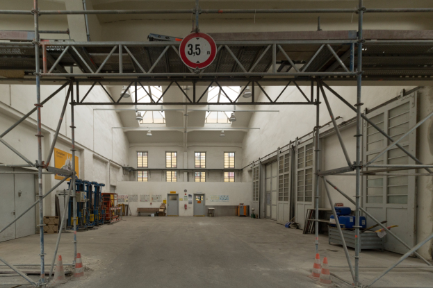 Großes Lagerhalle-Innere mit ausgedehnten Gerüsten, Verkehrskegeln, Metallstangen, einem Schild, Maschinen, einer Tür, Fenstern, Wänden, Deckenleuchten und Dach.