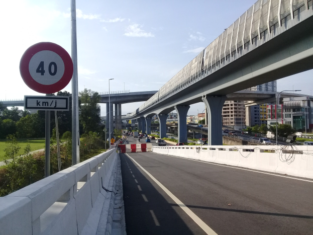 Straße mit Geschwindigkeitsbegrenzung, Fahrzeuge, Brücke mit Pfeilern, Laternen, Bäume, Gebäude und bewölkter Himmel im Hintergrund.