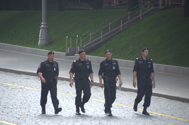 Vier Polizisten in Uniform gehen eine Straße entlang, jeder hält Papiere in der Hand, mit einem Pfahl, Treppe, Geländer, Gras, Bäumen und einem Gebäude im Hintergrund.