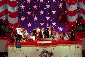 Eine Gruppe von Menschen steht auf einer Bühne mit einem Podium, das ein Logo trägt, umgeben von Luftballons und Konfetti, mit Sternen und einem Vorhang im Hintergrund.